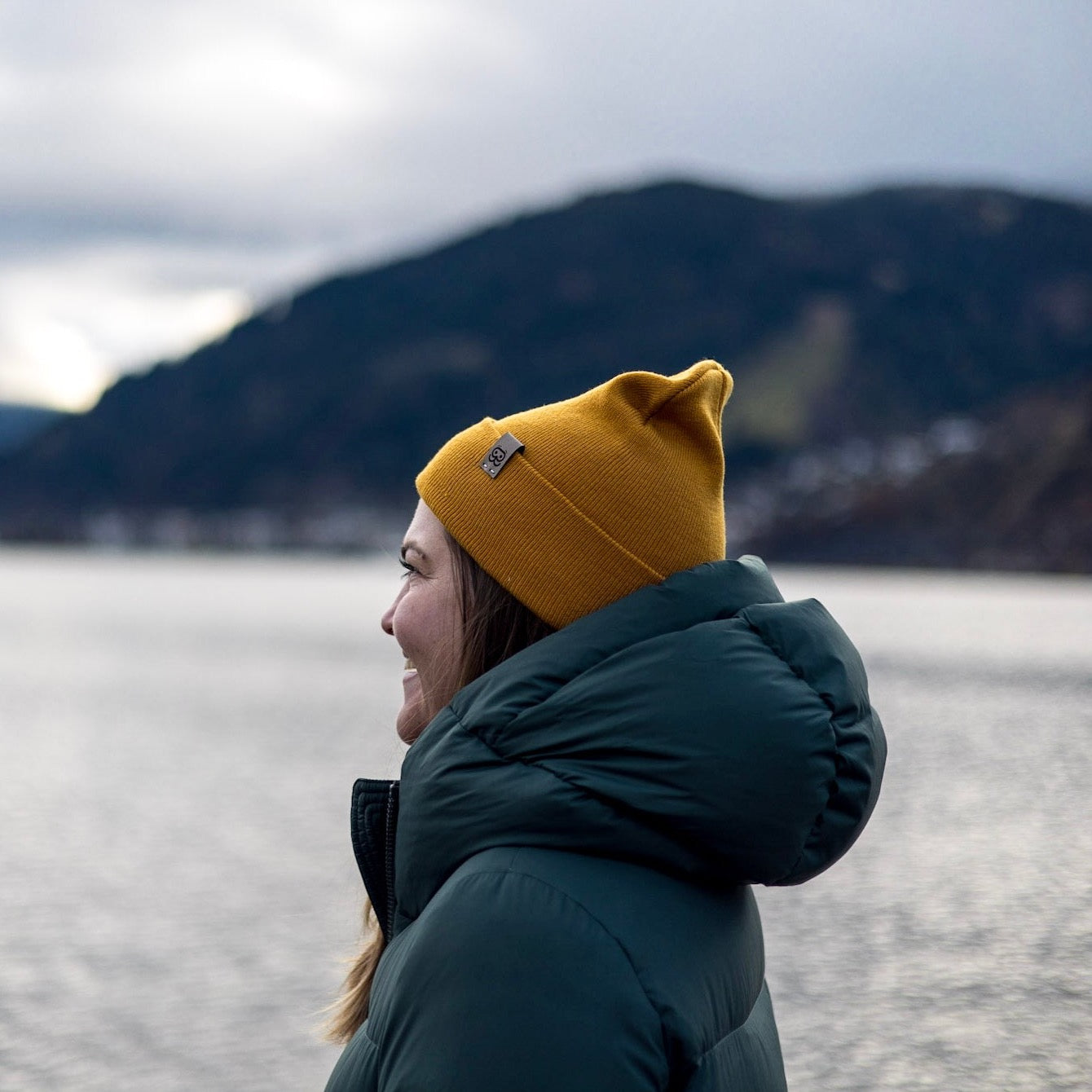 A person in a green puffy jacket made from recycled materials and a yellow Goggleface Color Beanie stands by a lake with mountains in the background. The cloudy sky suggests cold weather. The person is smiling, facing left, and enjoying the scenic view.