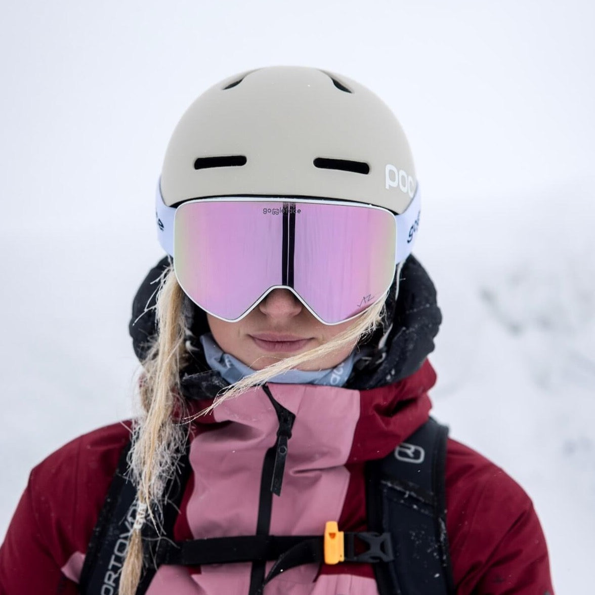 A person wearing the Goggleface Straight Line | White jacket and a helmet with pink-tinted snow goggles stands in a snowy environment. Strands of blonde hair are visible under the helmet, blending perfectly with the white, snowy background—a true Goggleface in winter wonderland.